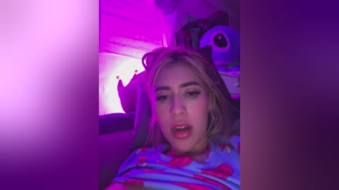 Video of a young woman with fair skin and blonde hair, wearing a colorful T-shirt, lying on a bed under purple lighting. She has a neutral expression, and a panda plush toy is in the background.