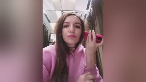 A video of a young woman with long brown hair, wearing a pink hoodie, taking a selfie in an airplane bathroom. She holds a red phone to her ear, reflecting her image in the mirror.