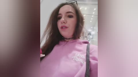 A video of a young woman with long brown hair, wearing a pink hoodie with \"I love things\" written on it. She has a black choker and is indoors, partially obscured by a blurred foreground.