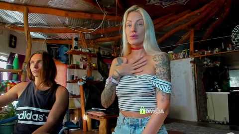 Video of a blonde woman with tattoos, wearing a striped crop top, standing in a cluttered, rustic room with a wooden ceiling, next to a brunette woman in a black Adidas tank top.