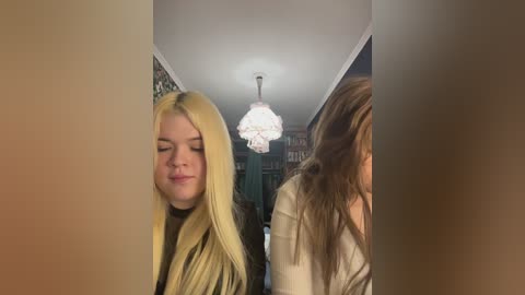 Video of two women with long blonde hair, one in the foreground smiling, the other in the background with a chandelier overhead, in a dimly lit room.