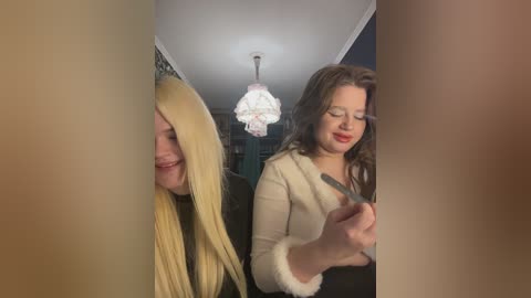 Video of two women: one with blonde hair, one with brunette hair, both in indoor setting, looking at phone, blurred background, chandelier.