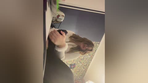A video captures a woman with long hair, wearing a white shirt and black pants, crouching in a bathroom with a floral-patterned shower curtain, holding a black phone.