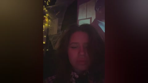 A dimly lit video of a young girl with long, wavy hair, wearing a dark jacket, gazing downward in a dimly lit room with fairy lights and a draped object in the background.
