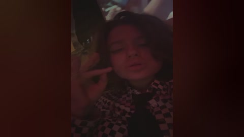 A video of a young person with long, wavy hair, wearing a black-and-white checkered shirt, lying on a bed, partially in shadow, holding a phone to their ear, looking tired.