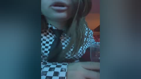 A video of a woman with light skin, wearing a black-and-white checkered shirt, mouth slightly open, blurred background, and a soft, muted color palette.