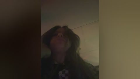 A dimly lit video of a woman with long dark hair, wearing a black shirt, stands in a room with beige walls and a darkened corner. Her expression is serious and her head is tilted upward.