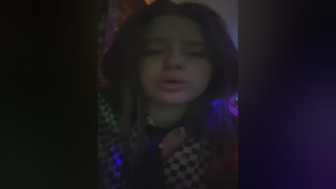 A blurry video shows a young girl with long, dark hair and closed eyes, wearing a patterned shirt, against a dimly lit, neon-lit background.