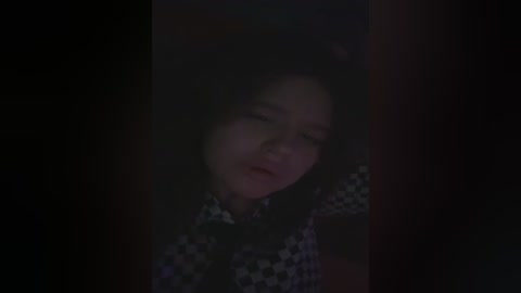 A dimly lit video captures a young girl with long dark hair, wearing a black and white checkered shirt, lying on a bed with her eyes closed, suggesting a somber or contemplative mood.