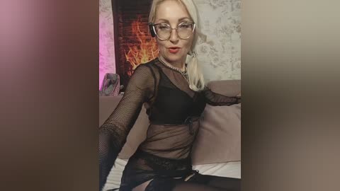 Media: Video of a fair-skinned, blonde woman with glasses and red lipstick, wearing black fishnet lingerie and stockings, sitting on a bed with a brick fireplace in the background.