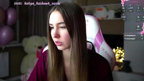 Media: A video of a young woman with long, straight brown hair, wearing a maroon shirt, sitting in a pink gaming chair in a softly lit room.