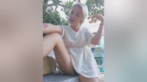 Video of a blonde woman in a sheer white dress, sitting on a balcony, legs crossed, with a tattoo on her arm, greenery in the background.