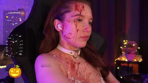 Video of a young woman with light skin and long brown hair, wearing a white choker and headphones, seated in a gaming chair, blood on her face, dimly lit room with Halloween decorations and a pumpkin.