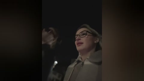 Video of a man with glasses, wearing a beige hoodie, looking up in dim lighting, possibly in a dark alley.