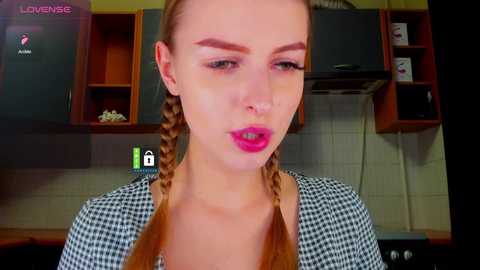 Video of a young Caucasian woman with light skin, pigtails, and bold pink lipstick, wearing a black-and-white checkered top, in a modern kitchen with wooden cabinets and white tiles.