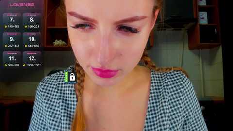 Video of a young woman with light skin and brown hair in braids, wearing a checkered blouse, looking down, in a kitchen with a \"Love Meter\" screen displaying stats on her face.