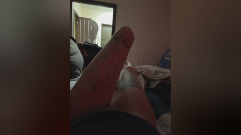 A dimly lit video of a person lying on a bed with their legs up, showing a tattooed penis. The room has a mirror reflecting a partially visible person and cluttered bedding.