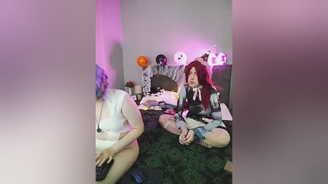 Video of two women dressed as anime characters, one with long red hair, sitting on a bed with green patterned sheets and Halloween decorations, in a dimly lit room.