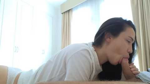 Video of a woman with fair skin and long dark hair performing oral sex on a man in a bright, softly lit room.