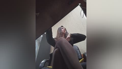 Media: A video of a young woman with long, straight hair, wearing a black sweater and dark tights, sitting on a chair, with a blurred, beige wall and ceiling in the background.