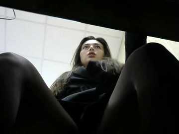 Media: Video of a young woman with long brown hair, wearing a black coat, seated in a car with legs spread, looking up. Background shows a white ceiling with fluorescent lights.