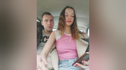 Video of a couple in a car, man with short hair, wearing a gray shirt, woman with wavy hair, pink tank top, beige jacket, blue jeans, both looking at a smartphone.