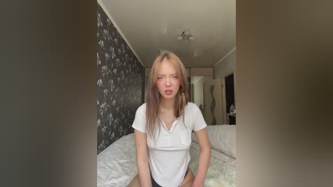 Media: Video of a young Caucasian woman with long, straight, light brown hair and fair skin, wearing a white t-shirt and standing in a bedroom with dark floral wallpaper and white bedding.