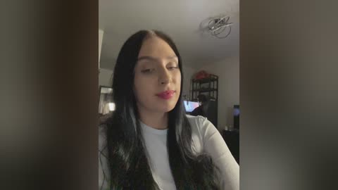 Video of a young woman with long black hair, fair skin, and red lipstick, wearing a white top and dark vest, standing indoors. Background shows a dimly lit living room with a TV and shelves.