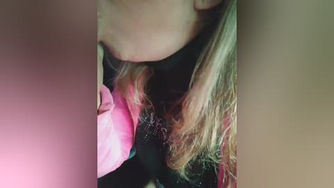 Media: Video of a person with light skin, blonde hair, and a black top, partially obscured by a pink jacket. The background is blurred and out of focus.