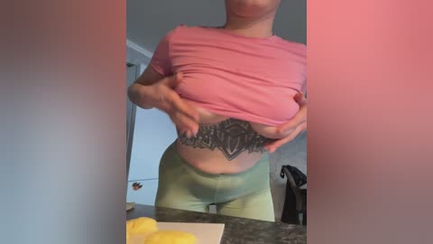 Video of a woman in a pink shirt lifting it to reveal a detailed black lace tattoo on her abdomen. She wears tight green pants. Background includes a kitchen counter with a yellow object and a door.