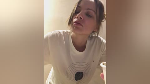 Video of a young woman with fair skin and brown hair, wearing a white t-shirt with a black geometric pattern, sitting in a dimly lit room with soft lighting.