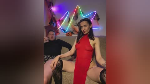 Video of a woman in a red, high-cut dress, seated provocatively with her legs spread, and a man in black pants and a black top, both smiling, in a room with neon lights and a Halloween theme.