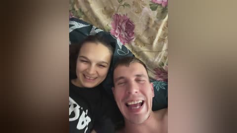Media: Video of a smiling young woman with curly hair, lying on a bed with a floral-patterned duvet, next to a shirtless man laughing, both appearing joyful.