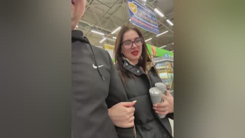 Media: Video of a middle-aged woman with glasses, wearing a black leather jacket, holding a gray coffee cup in a brightly lit supermarket aisle.