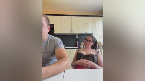 Media: A video of a man and woman in a modern kitchen. The man, with short hair, wears a gray t-shirt. The woman, with glasses and light skin, wears a black lace bra and red skirt, smiling.