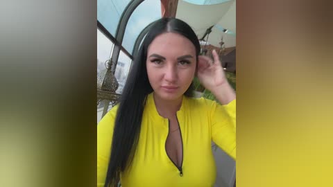 Video of a young woman with long black hair, wearing a bright yellow, long-sleeved top, adjusting her hair in a modern indoor setting with green plants and a chandelier.