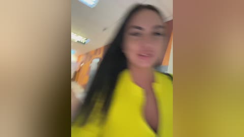 A blurry video of a young woman with long, dark hair, wearing a bright yellow blouse, taken indoors with a soft, warm light.