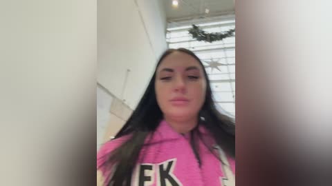 Video of a young woman with long black hair, wearing a pink hoodie with white text, standing indoors near a window with a Christmas wreath.