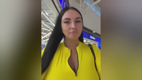A video of a young woman with long black hair, wearing a bright yellow zip-up hoodie, standing in a modern airport terminal with blue signage and white ceiling.