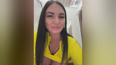 Video of a young woman with long black hair and fair skin, wearing a bright yellow top, smiling subtly, standing in a dimly lit room with beige walls and a window.