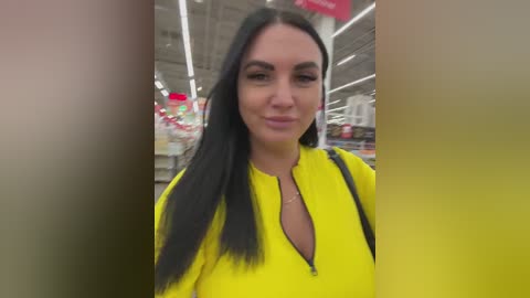 Video of a woman with long black hair, wearing a bright yellow top, standing in a grocery store aisle with shelves and products in the background.