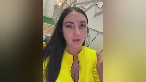 Video of a young woman with long black hair, wearing a bright yellow top, standing in a modern, industrial-style room with white walls and a staircase.