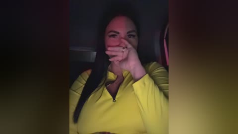 Video of a woman with long black hair, wearing a bright yellow zip-up jacket, covering her mouth with her hand, seated in a dimly lit car.
