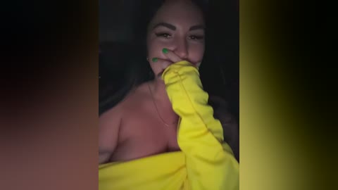 Video of a woman with olive skin, long dark hair, wearing a yellow shirt, holding it to her mouth, looking seductively at the camera, with a blurred background.