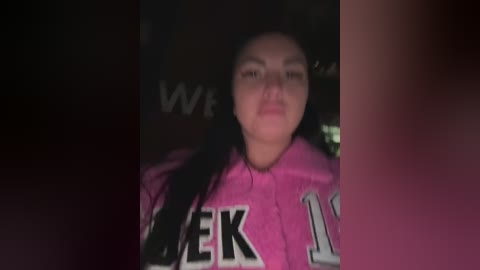 Video of a young woman with long black hair wearing a pink hoodie with large white lettering, likely \"WELCOME.\" The background is dark and indistinct, creating a moody, low-light atmosphere.