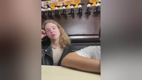 Media: Video of a young woman with shoulder-length blonde hair, wearing a black leather jacket over a white shirt, lounging on a counter, with a backdrop of a drink dispenser and boxes.