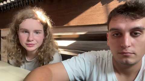 Media: Video of a young Caucasian girl with curly blonde hair and a fair complexion, smiling, next to a young man with short dark hair, wearing a white T-shirt, in a wooden room with sunlight streaming through the blinds.