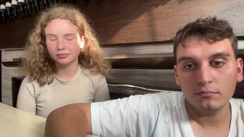 Media: A video of a young man with short dark hair in a white t-shirt, looking downcast, and a curly-haired girl in a beige top, smiling, in a cozy, wooden room with wine bottles on a shelf in the background.