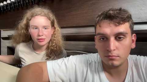 Media: Video of a young woman with curly blonde hair, wearing a gray shirt, and a young man with short brown hair, wearing a white shirt, in a bedroom with a wooden headboard and black coat hooks.