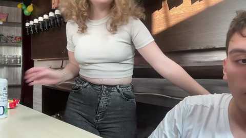 Media: Video of a blonde woman in a white crop top and high-waisted jeans, gesturing at a young man in a white T-shirt, seated at a counter in a dimly lit, rustic-themed bar.